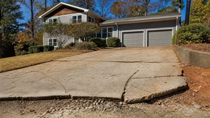 A worker examines an old driveway before replacement. at a grey split-level home in Marietta, GA, in clear late-morning sun.